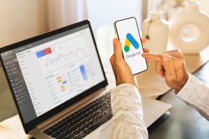 Man using a mobile phone with Google Ads on the screen and in the background a computer showing ad statistics. Rosario, Argentina - March 23, 2024